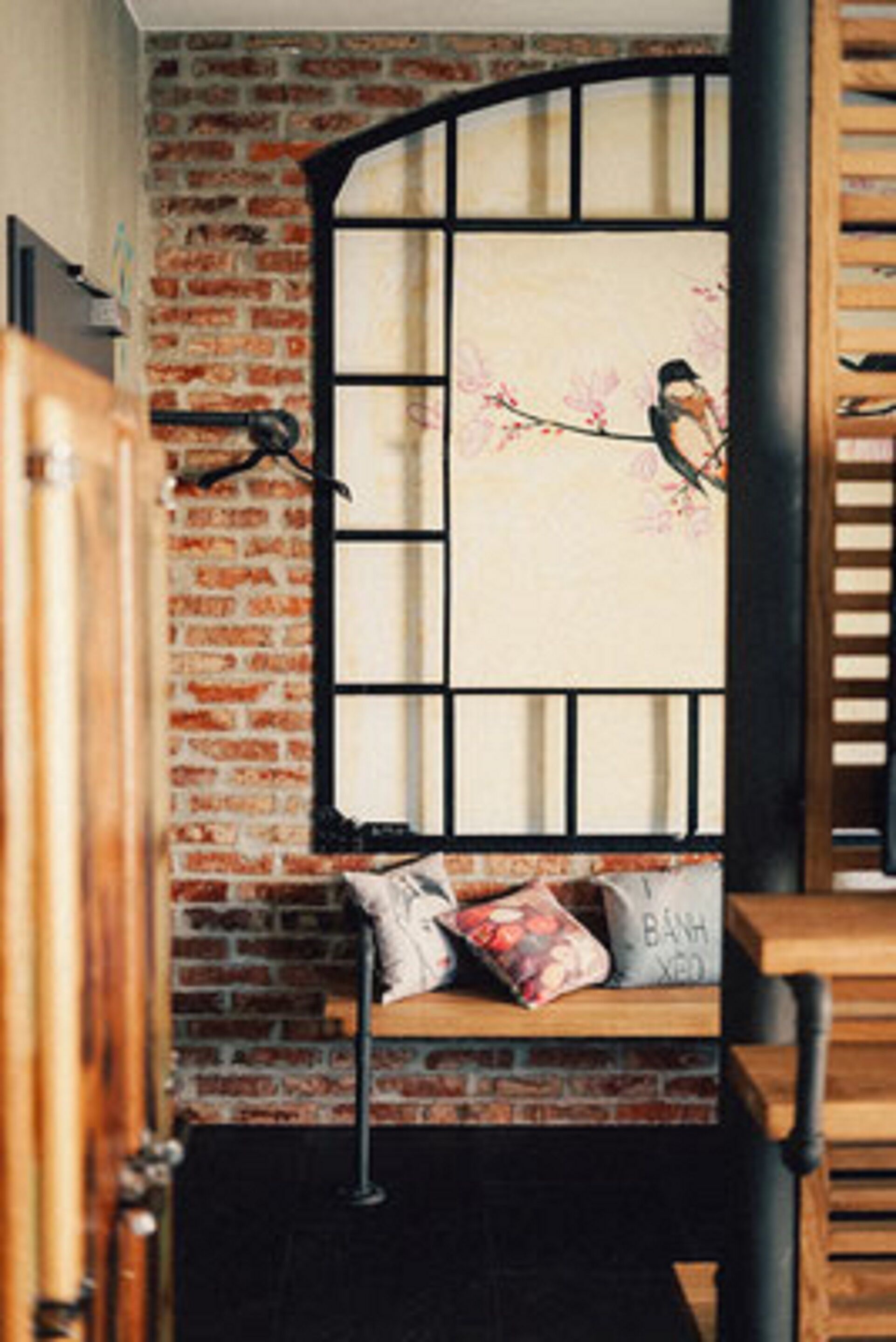 A room with a brick wall, a window with a black frame, a bench with cushions, and a wooden chair.