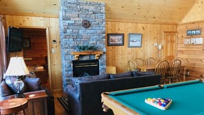 A cozy cabin interior with a stone fireplace, a pool table, a sofa, and wooden walls.