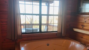 A wooden cabin room with a large bathtub, a window with curtains, and a wooden deck visible through the window.
