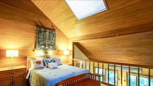 A bedroom with a wooden ceiling, a bed with a bench, a nightstand, a lamp, and a view through a window.