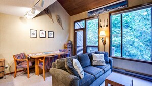 A cozy living room with a sofa, chairs, a dining table, and a large window offering a view of trees.