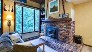 A cozy living room with a brick fireplace, a large window with a view of trees, a sofa with cushions, and a lamp.