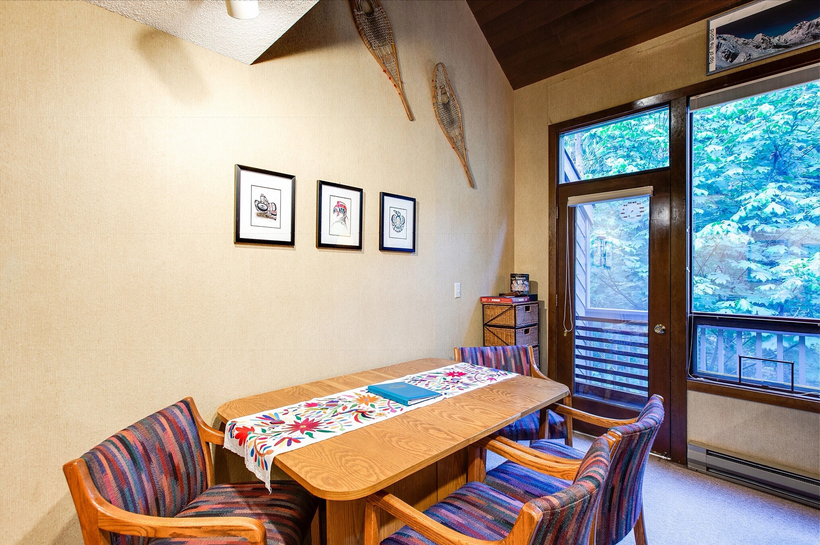 Un comedor con una mesa de madera y sillas a rayas, cuadros en la pared y vista a árboles a través de un ventanal grande.
