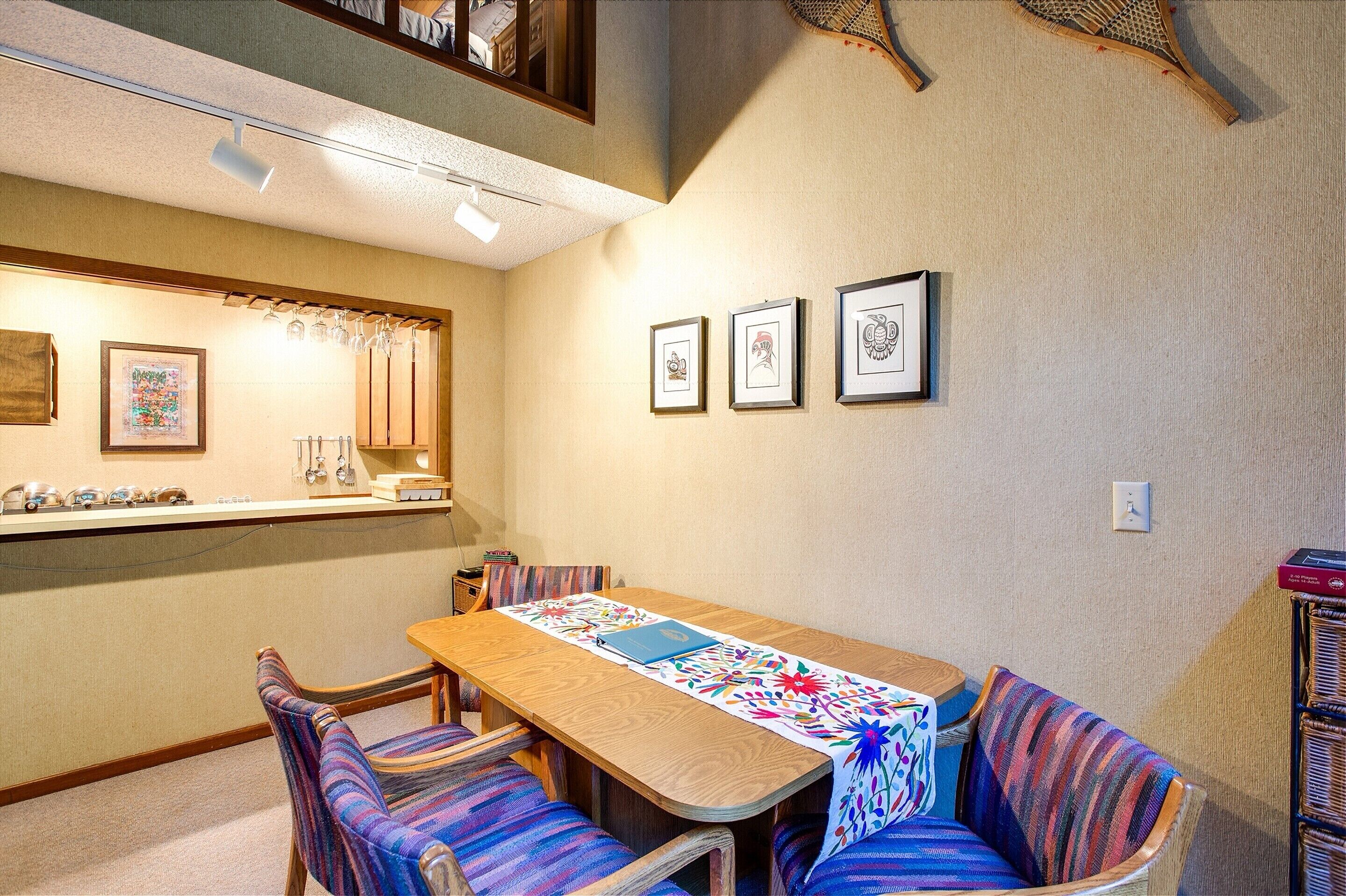 A dining area with a wooden table, striped chairs, and a colorful table runner. There are framed pictures on the wall and a shelf with decorative items.