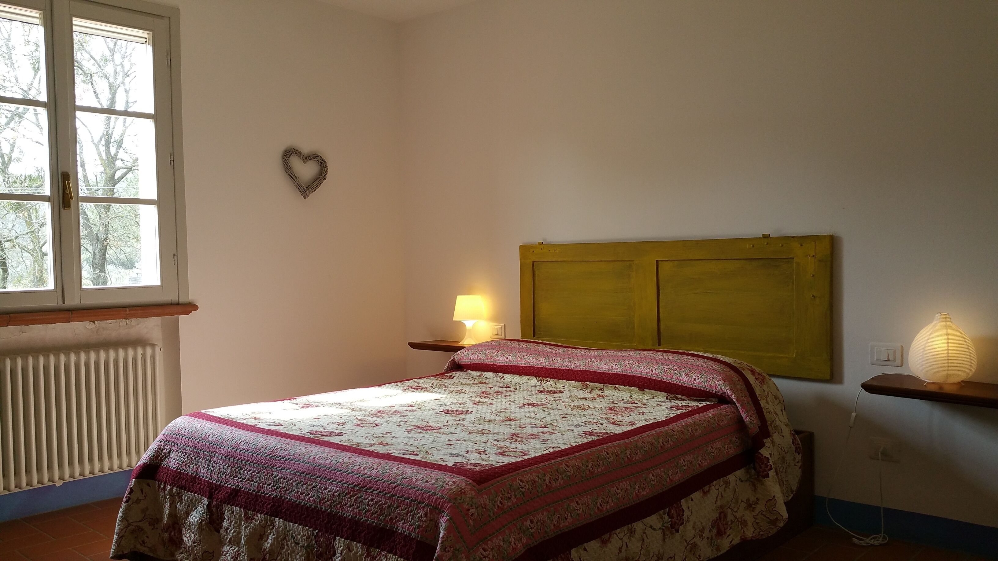 A bedroom with a bed, a nightstand with a lamp, a radiator, a window with a view of trees, and a heart-shaped decoration on the wall.