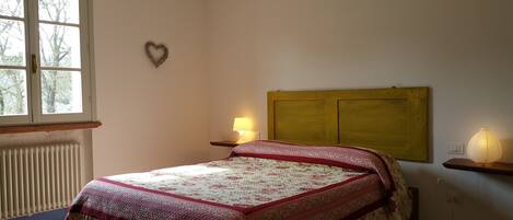 A bedroom with a bed, a nightstand with a lamp, a radiator, a window with a view of trees, and a heart-shaped decoration on the wall.