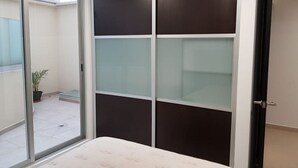 Une chambre avec une porte-fenêtre coulissante donnant sur un balcon, un lit avec une tête de lit blanche et une armoire de couleur sombre dotée de panneaux en verre dépoli.