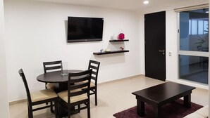 Un salon moderne avec une table à manger ronde, quatre chaises, un téléviseur à écran plat fixé au mur et une porte-fenêtre coulissante donnant sur un balcon.