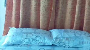 Uma cama com colcha estampada e duas almofadas, cortina com padrão floral e um interruptor de parede.