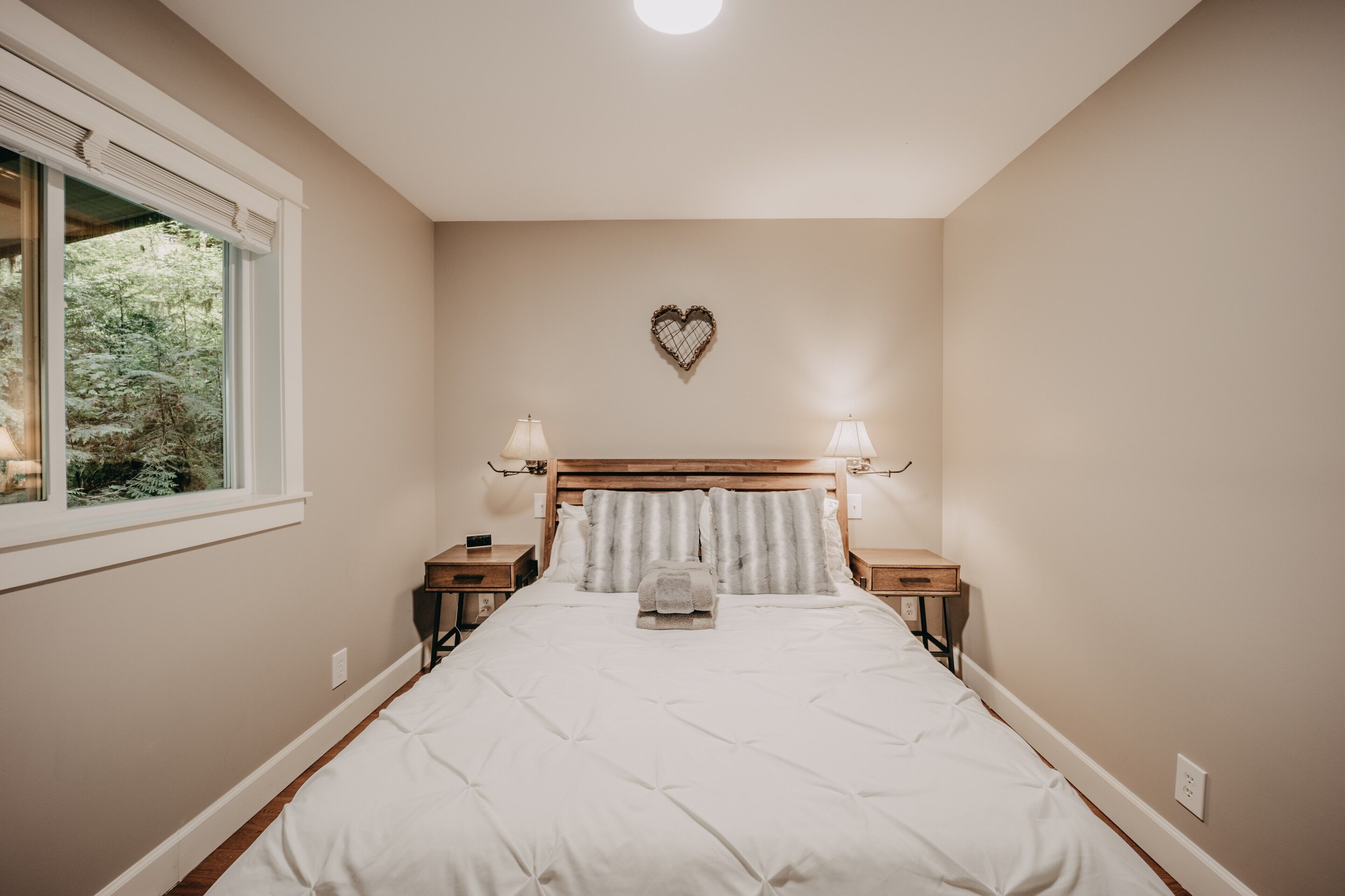 A bedroom with a large bed, two bedside tables, a window, and a heart-shaped wall decoration.