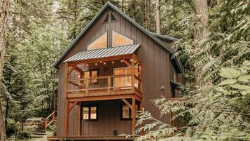 Una cabaña con terraza de madera y balcón, rodeada de un denso bosque.