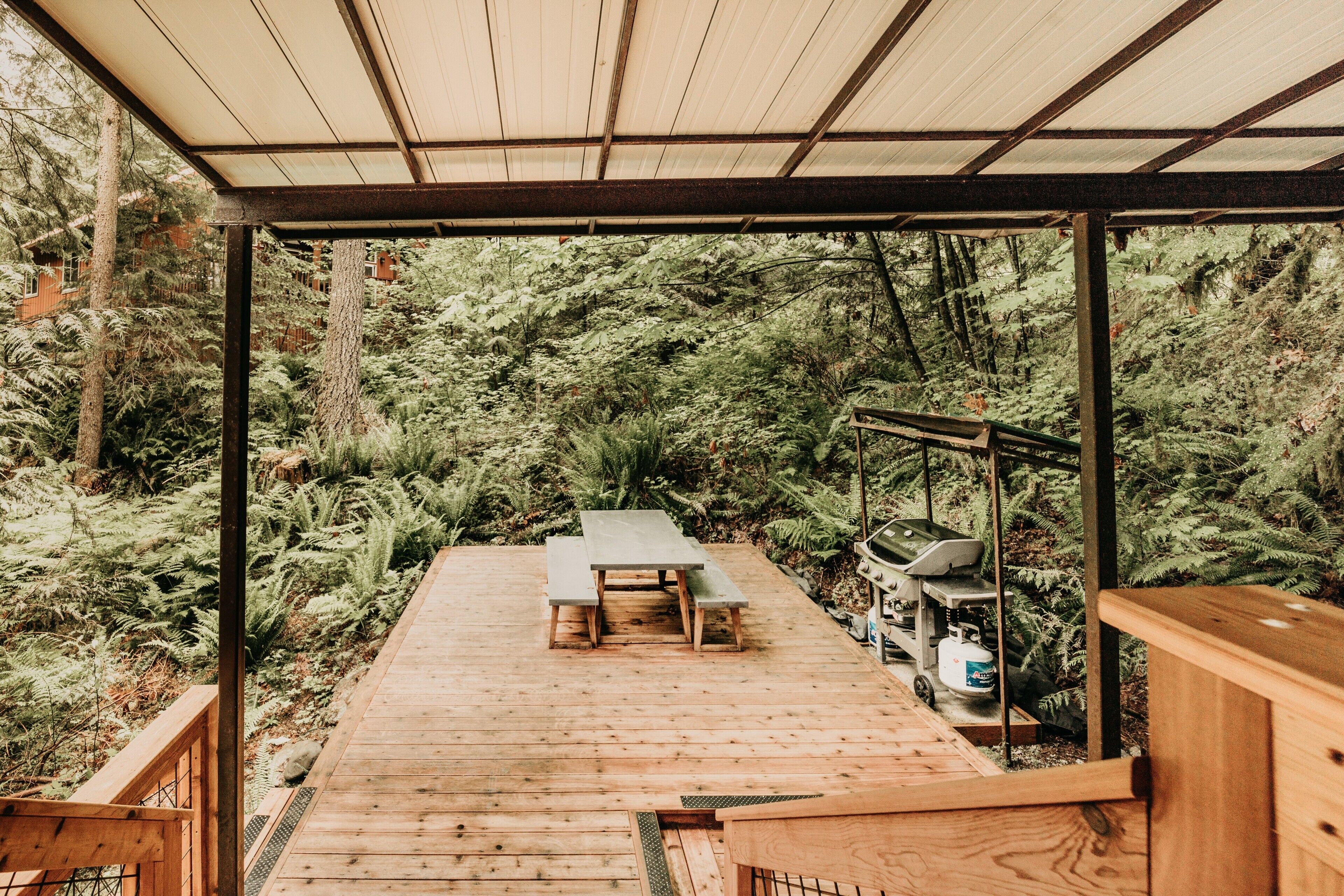Una terraza de madera con una mesa de picnic y zona de barbacoa, rodeada de un bosque denso.