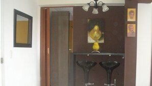 Un pasillo con suelo de baldosas, una puerta, un espejo, una araña y una pequeña mesa con un florero.