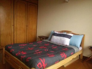 Quarto com cama, guarda-roupa de madeira e pequena mesa de cabeceira.