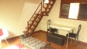 A room with a wooden staircase, a dining table with chairs, a patterned sofa, and a small table with a chair.