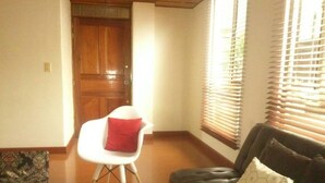 A living room with a wooden door, a white chair with a red cushion, a black leather sofa with patterned cushions, and a window with blinds.