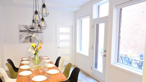 A dining area with a wooden table set for six, surrounded by black and white chairs. A vase with flowers is on the table, and there is a painting on the wall above it. - FALL & WINTER SPECIAL! Huge Ground Floor apt with Courtyard - 5 bed + 6 bath (Montréal)