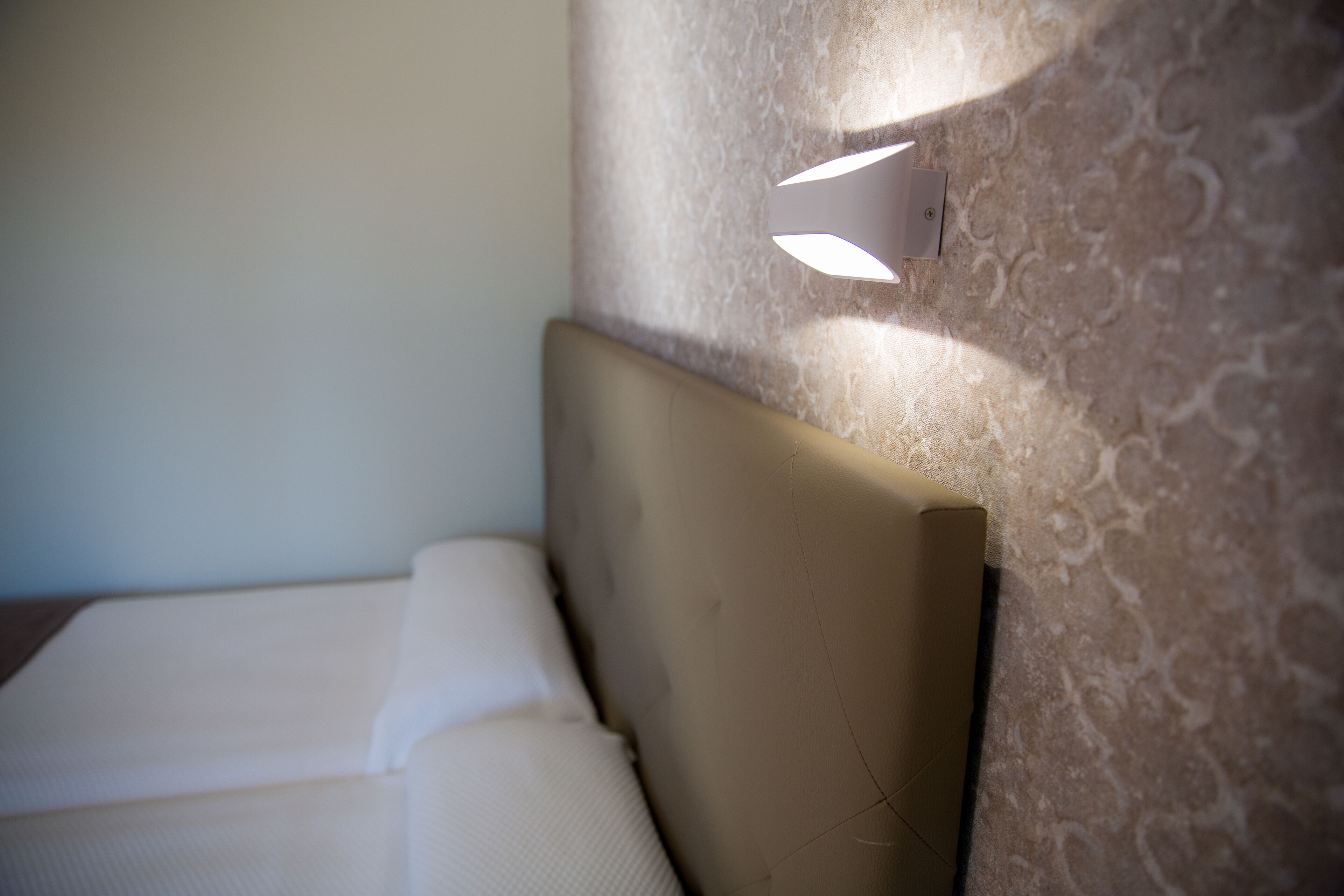 Een net opgemaakt bed met hoofdbord tegen een structuurwand, een wandlamp en een effen muur.