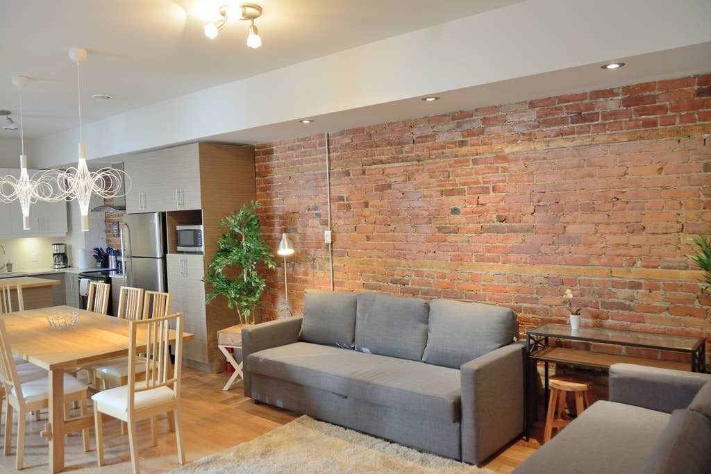 A modern living room with a grey sofa, wooden dining table, and chairs. A brick wall and a kitchen with a refrigerator and microwave are visible in the background.