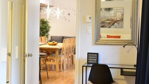 Eine offene Tür führt in einen Essbereich mit einem Holztisch, Stühlen und einer modernen Kronleuchter. Ein gerahmtes Kunstwerk hängt an der Wand, und ein Schreibtisch mit Stuhl ist im Vordergrund zu sehen.