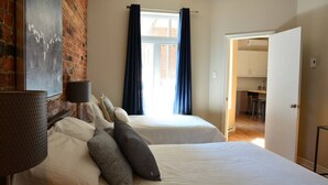 Ein Schlafzimmer mit zwei Betten, einer Ziegelwand, einem Fenster mit Vorhängen und einer Tür zu einer Küche.
