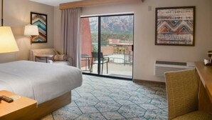 A hotel room with a bed, a desk, a chair, a table, a lamp, a window with a view of mountains, and artwork on the walls.