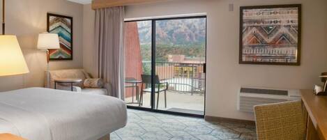 Habitación de hotel con cama, escritorio, silla, mesa, lámpara, ventana con vistas a montañas y obras de arte en las paredes.