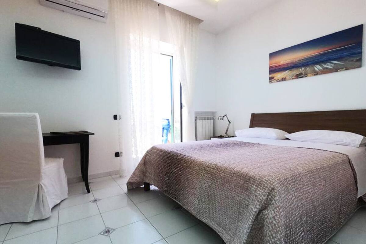 A hotel room with a bed, a desk, a chair, a TV, and a painting on the wall.
