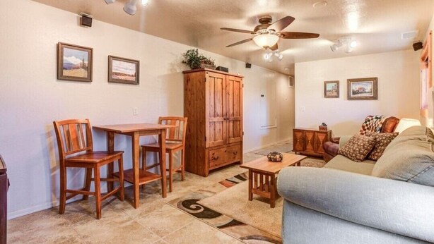 A living room with a wooden dining table, two chairs, a sofa, a ceiling fan, and framed pictures on the walls.