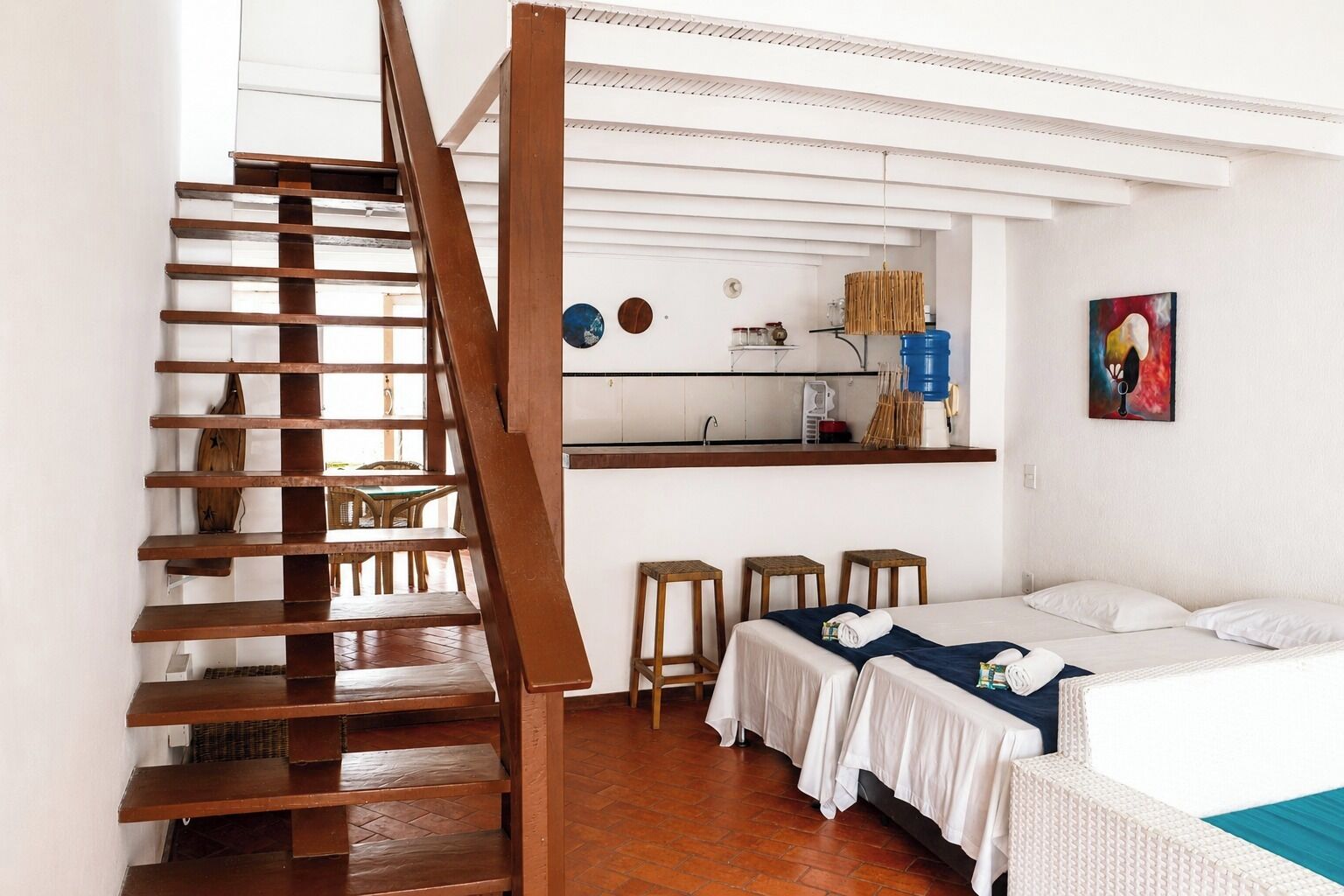A room with a wooden staircase, a bed with white bedding, a small kitchen area, and a painting on the wall.