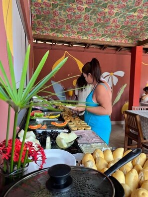 Uma mulher servindo comida em um buffet ao ar livre, com diversos pratos e uma churrasqueira em primeiro plano.