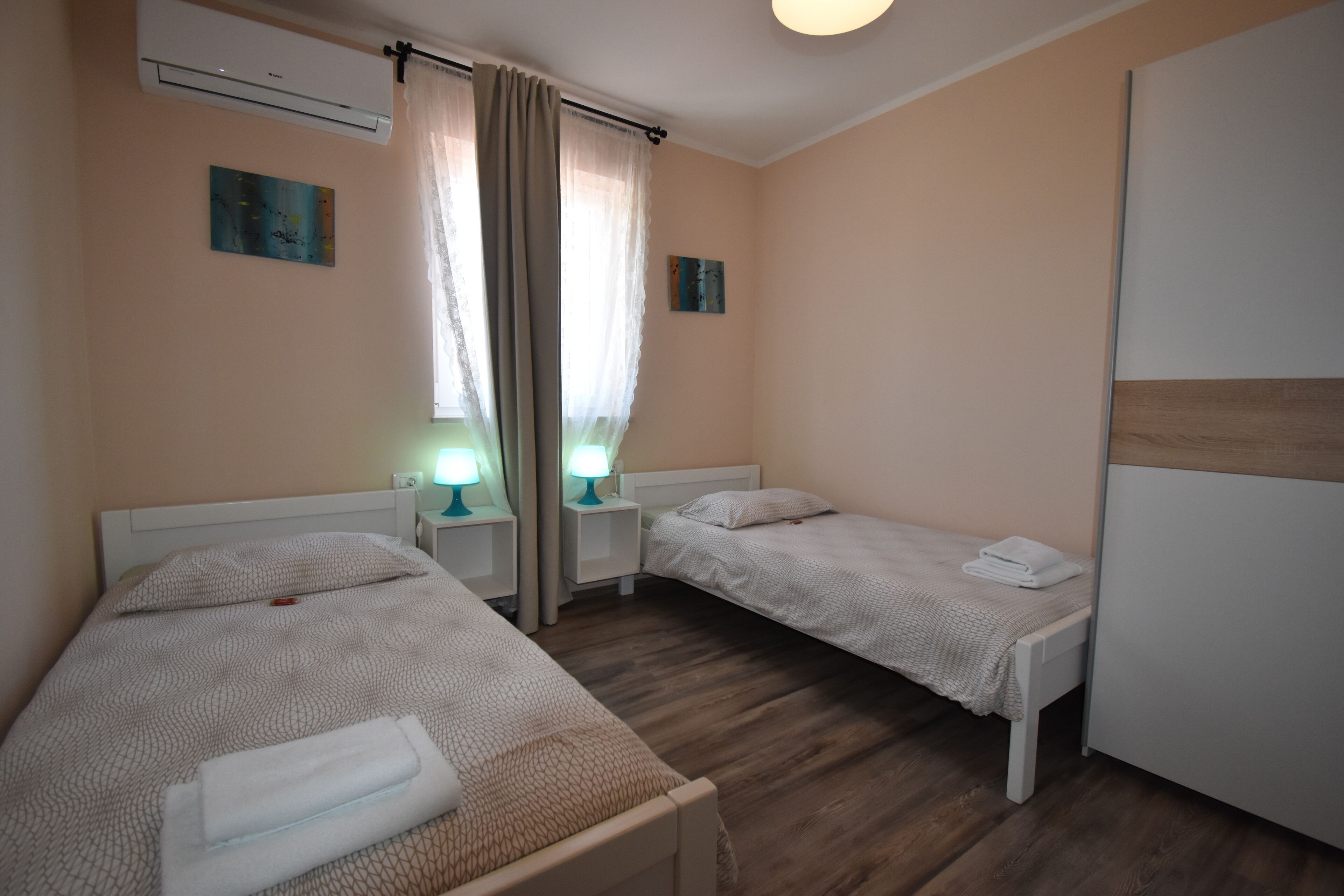 Habitación de hotel con dos camas individuales, un armario, una mesita de noche con lámpara y una ventana con cortinas.