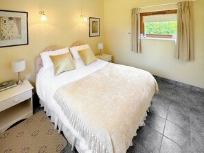A neatly made bed with a white and beige blanket, two bedside tables with lamps, a framed picture on the wall, and a window with curtains. - Lovely single-storey cottage with garden in a quiet rural location. (Hassocks)