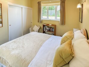 A bedroom with a bed, a chair, a bookshelf, and a window with curtains. - Lovely single-storey cottage with garden in a quiet rural location. (Hassocks)