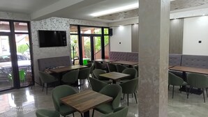Interior de un restaurante con varias mesas y sillas, un televisor en la pared y amplios ventanales que aportan luz natural.