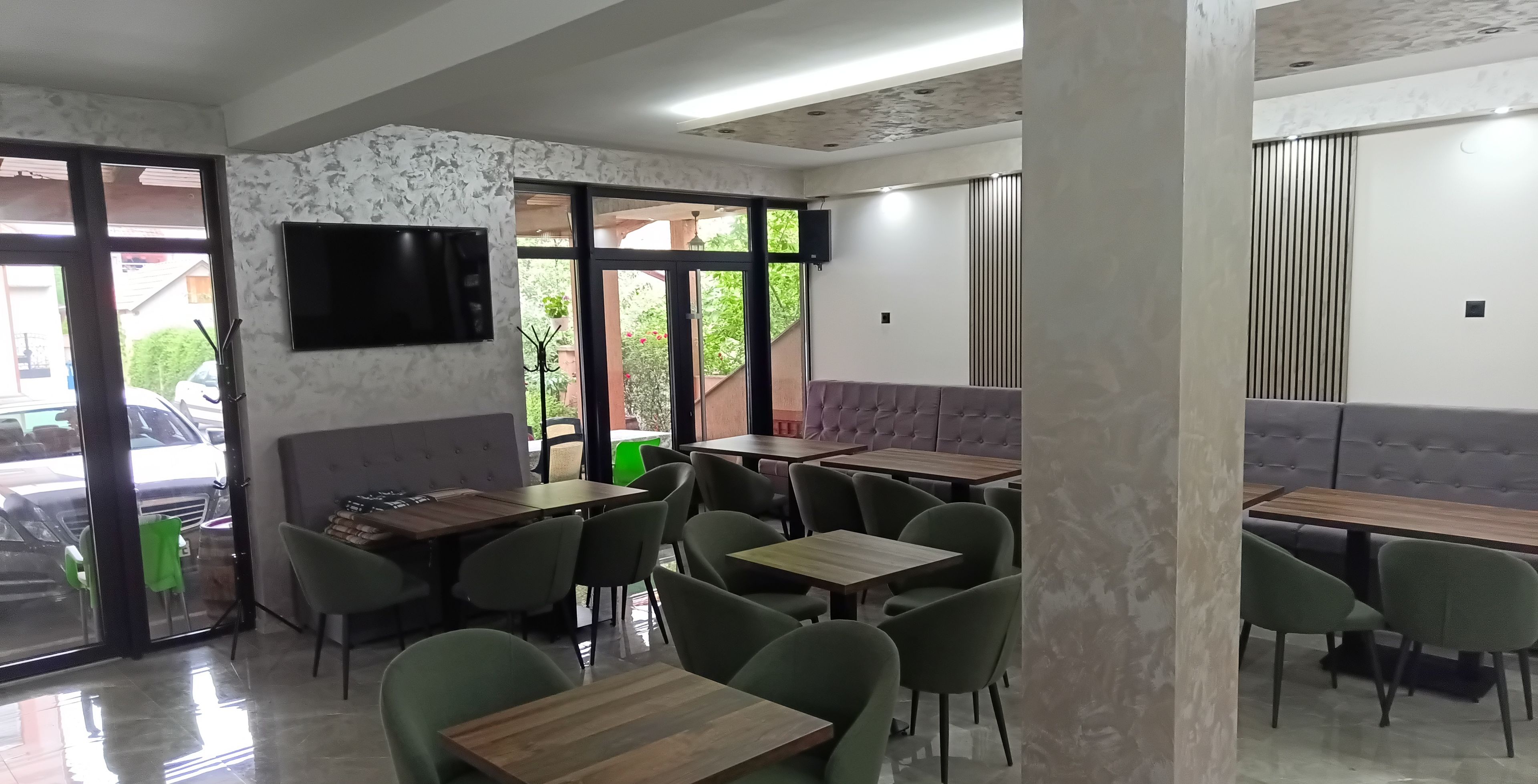 A restaurant interior with multiple tables and chairs, a television mounted on the wall, and large windows providing natural light.