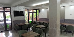 Interior de un restaurante con varias mesas y sillas, un televisor en la pared y amplios ventanales que aportan luz natural.
