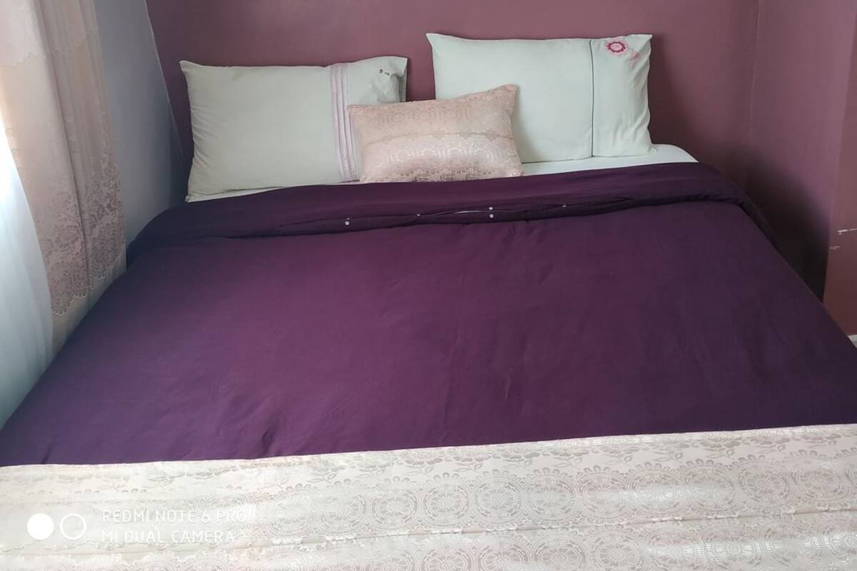 Cama com colcha roxa e duas almofadas, uma verde clara e outra bege, num quarto com paredes roxas.