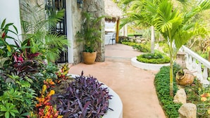 A tropical garden pathway leading to a building with a stone facade and a thatched roof.