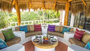 A circular seating area with a central coffee table, surrounded by cushioned sofas with colorful pillows, set against a backdrop of lush greenery and a thatched roof structure.