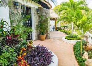 A tropical garden pathway leading to a building with a stone facade and a thatched roof.