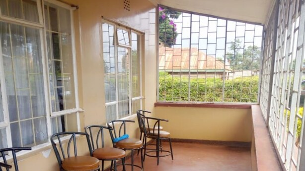 A covered outdoor seating area with four chairs and a table, large windows with metal bars, and a view of greenery and buildings in the background. - Banana Leaf Hotel (Nyeri)