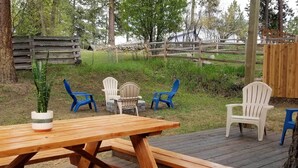 Un muelle de madera con una mesa de picnic y cuatro sillas Adirondack, rodeado de un cerco de madera y follaje.