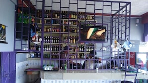 A bar with shelves stocked with various bottles, a television mounted above the bar, and a Coca-Cola refrigerator in the background. - Millennium Guest House (Narok)