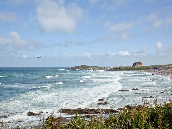 A coastal landscape with waves, a beach, and a large building in the distance. - Apartment 2 Waters Edge (Newquay)