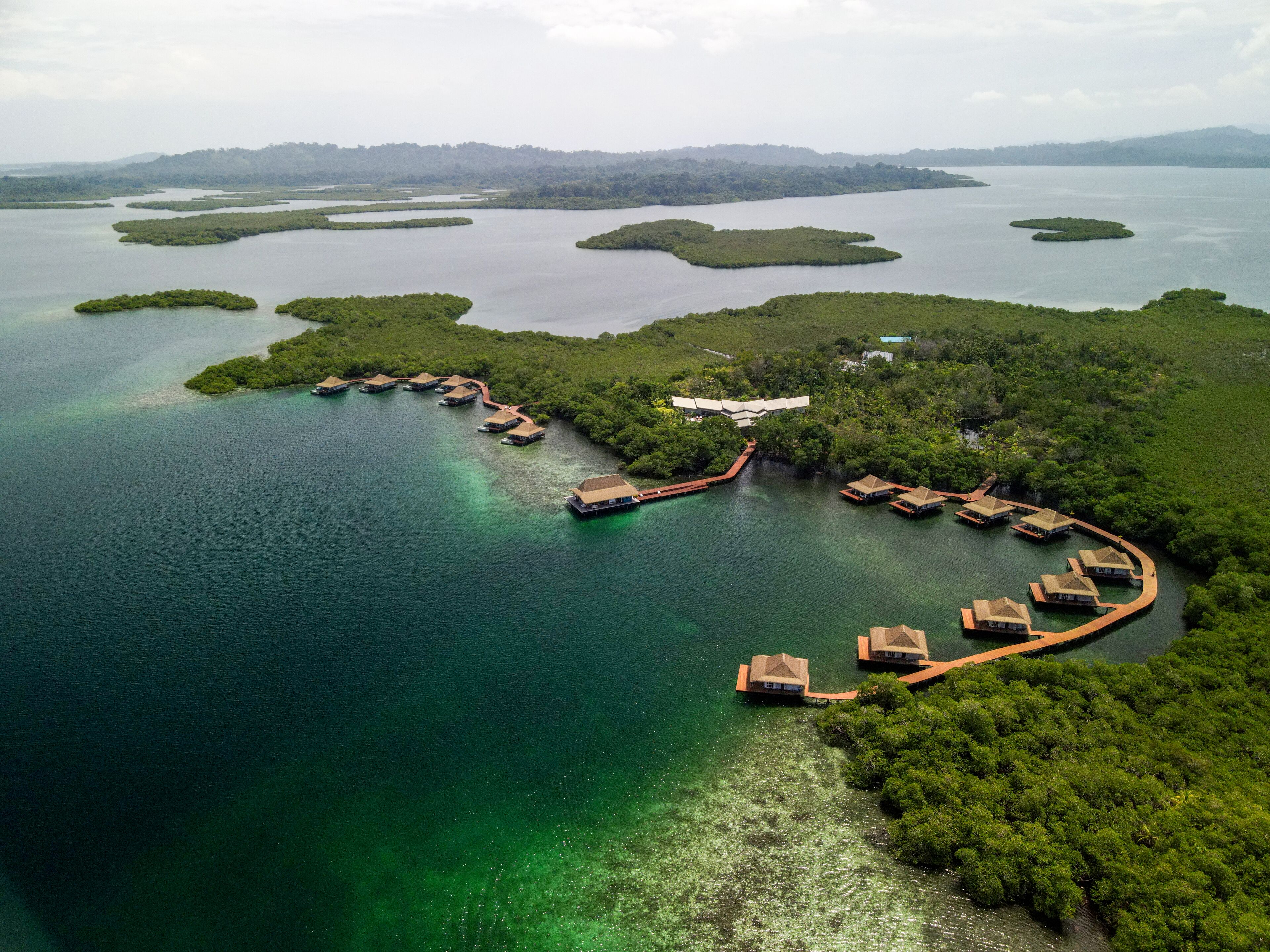 An aerial view of a resort with overwater bungalows connected by walkways, surrounded by clear blue waters and lush greenery.