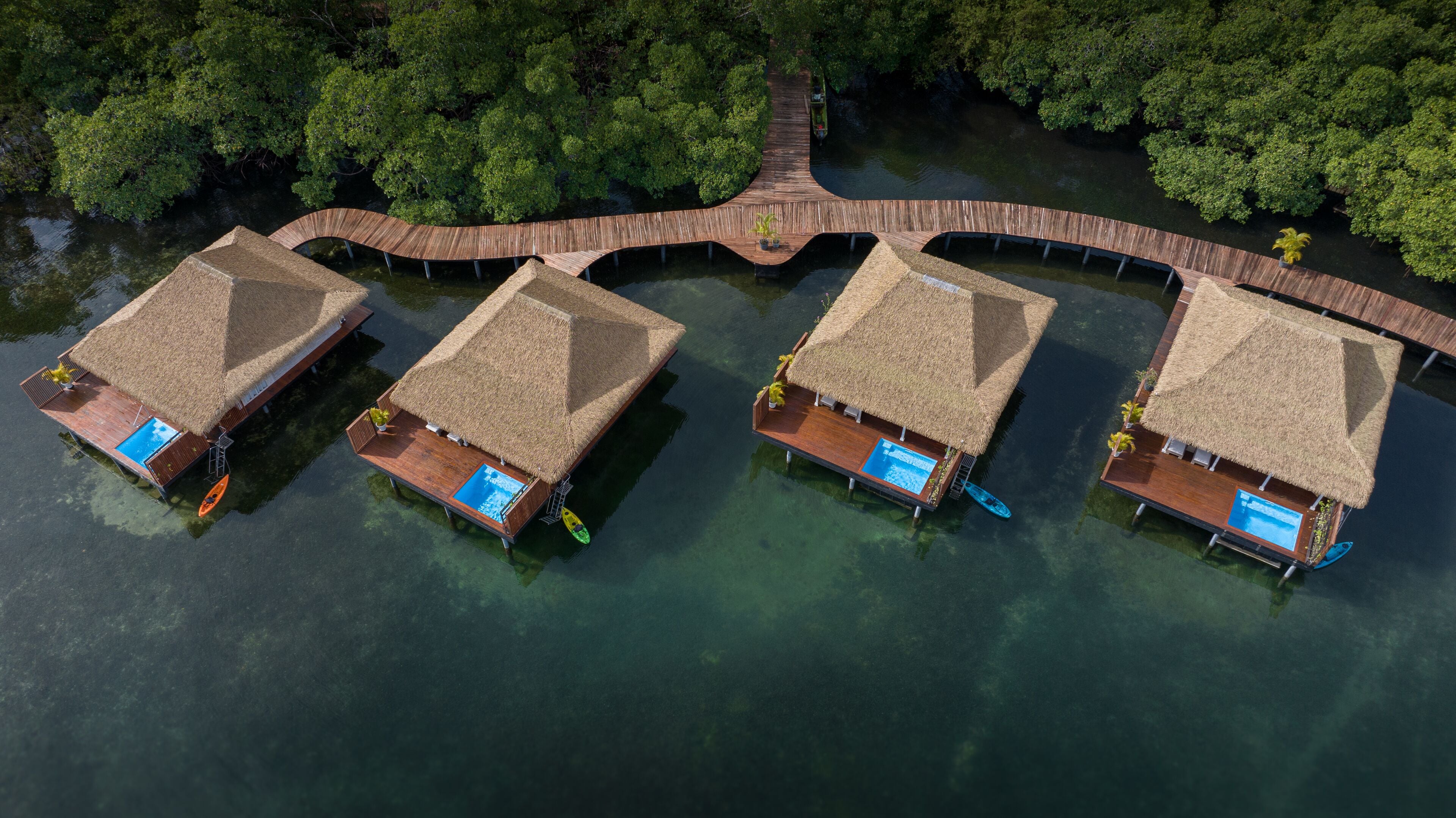 Vista aérea de un resort flotante con bungalows sobre el agua con techo de paja, unidos por un pasillo de madera. Cada bungalow tiene una piscina y está rodeado de agua.