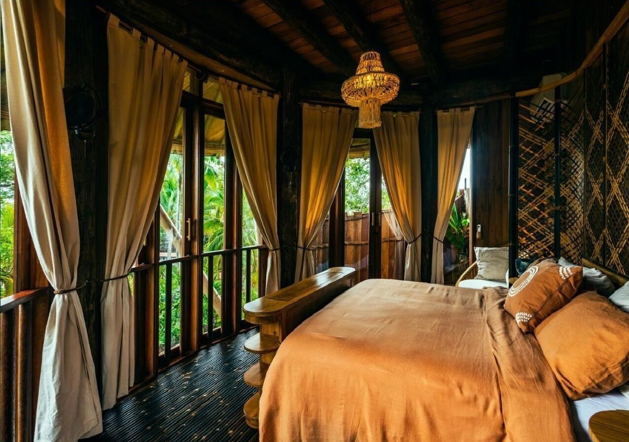 A bedroom with a bed, curtains, a wooden bench, and a hanging light fixture.