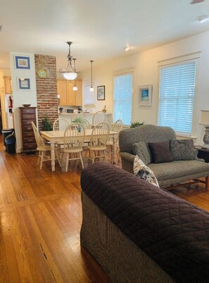A cozy living room with a dark-colored sofa and a matching ottoman, a dining area with wooden chairs and a table, and a kitchen with wooden cabinets and a brick accent wall.