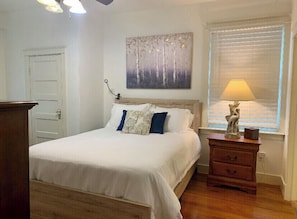 A bedroom with a bed, a nightstand, a lamp, a painting, and a window with blinds.